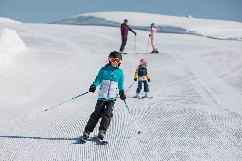 Dzieci na nartach na stoku, w tle dwie dorosłe osoby