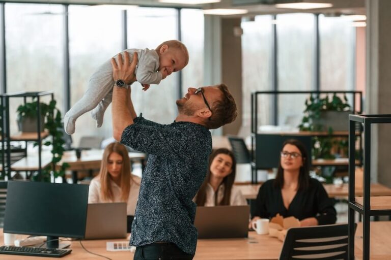 Mężczyzna podnoszący dziecko w biurze, w tle 3 kobiety pracujące przy komputerze