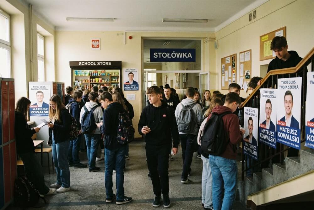 Uczniowie mijają w korytarzu szkolnym plakaty wyborcze rozwieszone w strategicznych miejscach przy sklepiku, stołówce i schodach.