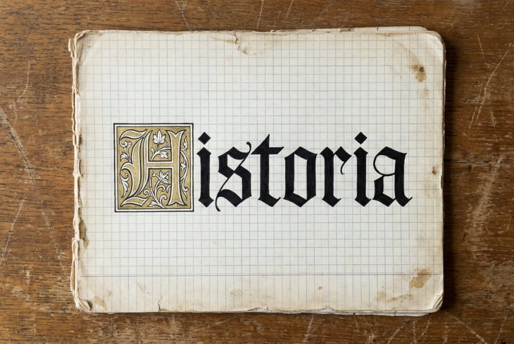 Pierwsza strona zeszytu z gotyckim napisem „HISTORIA” i ozdobnym złotym inicjałem na drewnianym biurku.